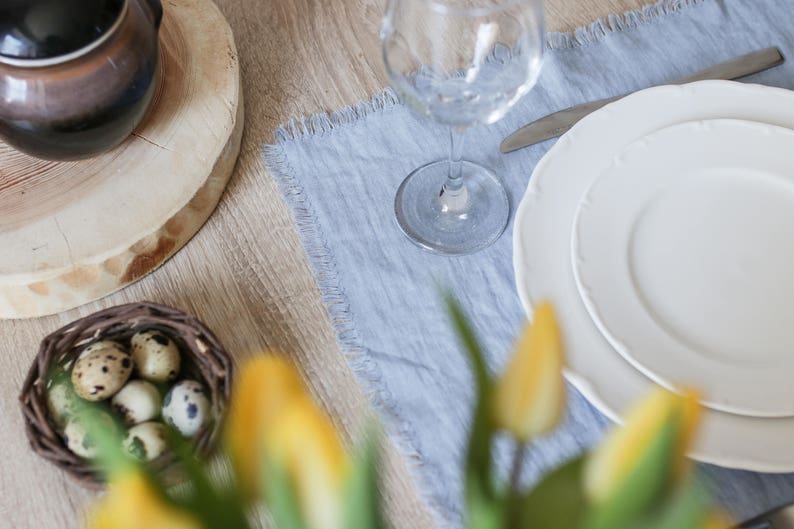 Pu&ograve; includere: Un tovaglietta di lino blu con un piatto bianco e un bicchiere d'acqua. In primo piano ci sono dei tulipani gialli e sullo sfondo una ciotola di uova di quaglia.