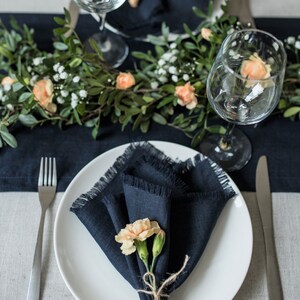 May include: A white dinner plate with a navy blue linen napkin folded into a triangle and topped with a small bouquet of peach-colored flowers. The plate is set on a gray linen tablecloth with a navy blue linen runner and a silver fork and knife.