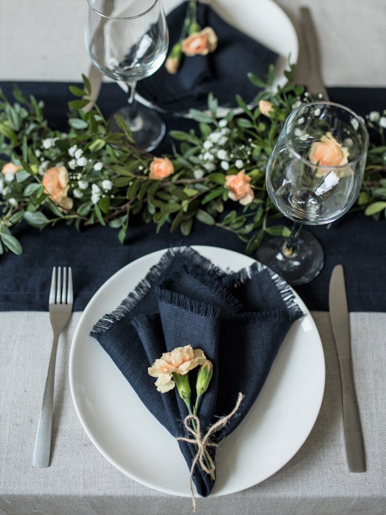 May include: A white dinner plate with a navy blue linen napkin folded into a triangle and topped with a small bouquet of peach-colored flowers. The plate is set on a gray linen tablecloth with a navy blue linen runner and a silver fork and knife.