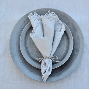 May include: A gray ceramic plate with a smaller gray ceramic plate inside. A white linen napkin with frayed edges is placed on top of the plates.