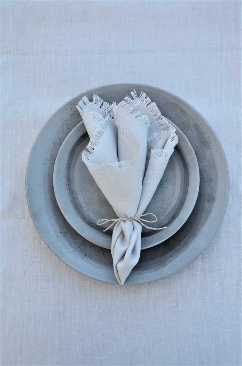 May include: A gray ceramic plate with a smaller gray ceramic plate inside. A white linen napkin with frayed edges is placed on top of the plates.