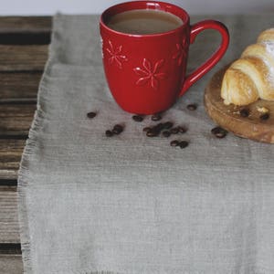 May include: A red mug with a snowflake design filled with coffee sits on a gray linen tablecloth with coffee beans scattered around it. A croissant is on a wooden cutting board in the background.