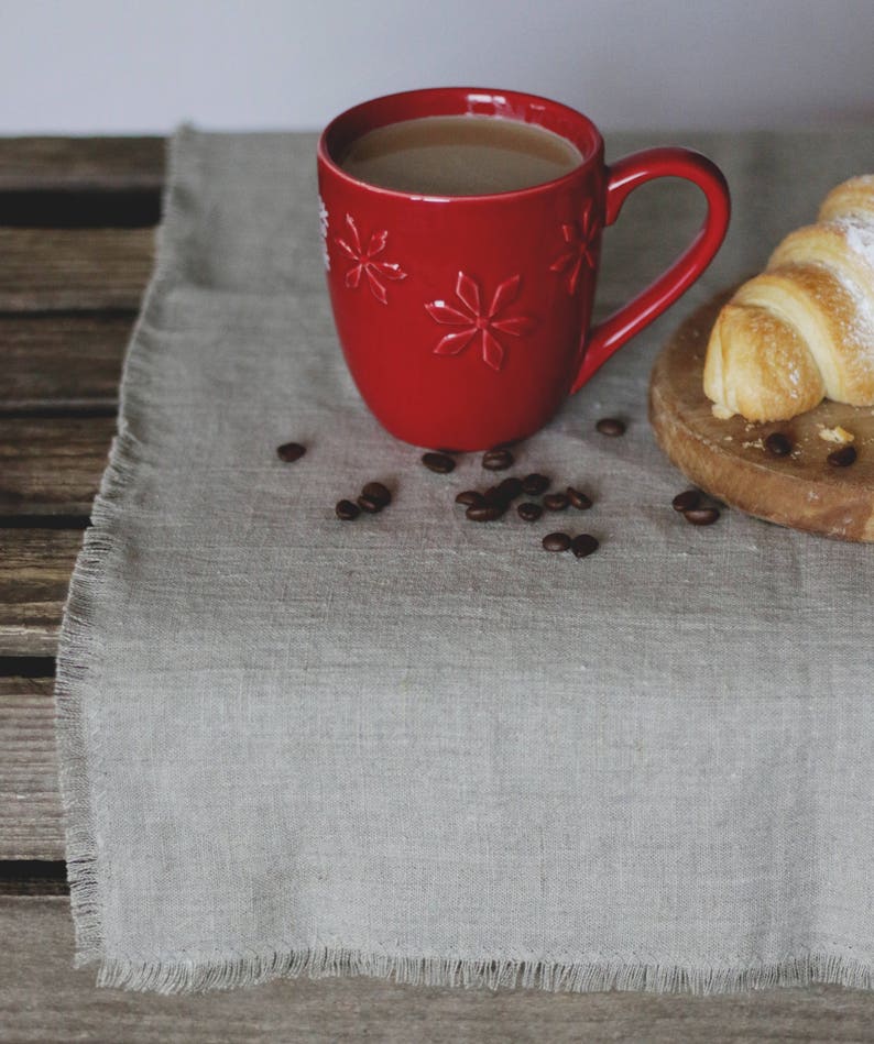 May include: A red mug with a snowflake design filled with coffee sits on a gray linen tablecloth with coffee beans scattered around it. A croissant is on a wooden cutting board in the background.