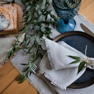 Pu&ograve; includere: Un tovagliolo di lino grigio con bordi sfrangiati &egrave; piegato e legato con uno spago, adornato con un rametto di verde. Il tovagliolo &egrave; posto su un piatto in ceramica blu scuro, che &egrave; appoggiato su un sottopiatto di lino bianco. Una forchetta e un coltello d'argento sono anche visibili sul sottopiatto.