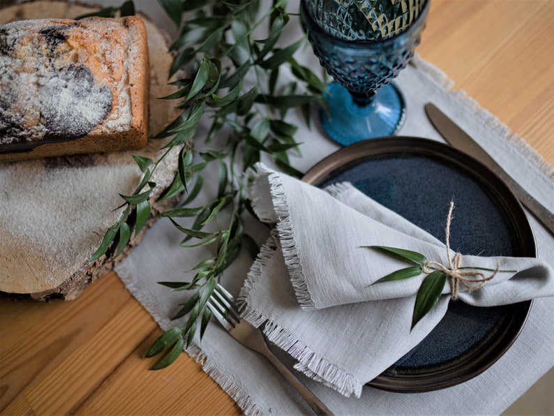 Pu&ograve; includere: Un tovagliolo di lino grigio con bordi sfrangiati &egrave; piegato e legato con uno spago, adornato con un rametto di verde. Il tovagliolo &egrave; posto su un piatto in ceramica blu scuro, che &egrave; appoggiato su un sottopiatto di lino bianco. Una forchetta e un coltello d'argento sono anche visibili sul sottopiatto.