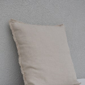 May include: A beige linen pillow with a frayed edge. The pillow is sitting on a white surface.