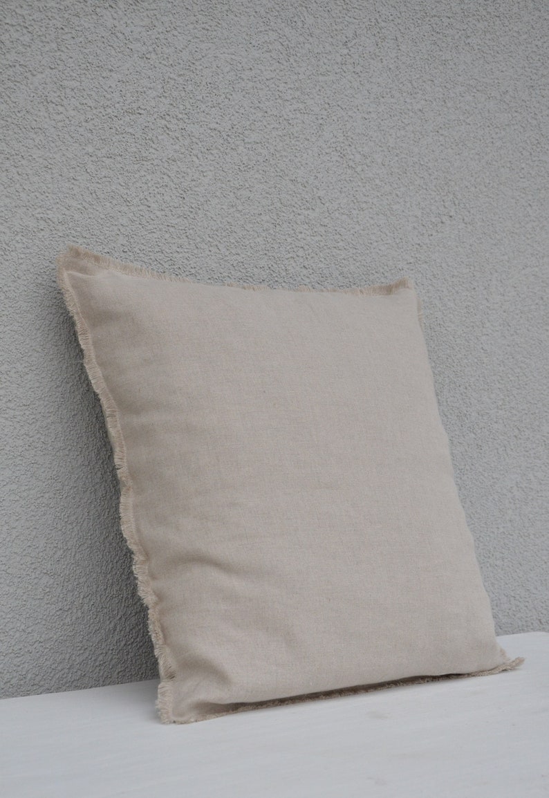 May include: A beige linen pillow with a frayed edge. The pillow is sitting on a white surface.