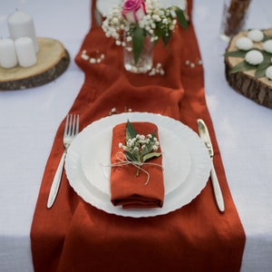 May include: A white table setting with a rust-colored linen runner. A white plate with a rust-colored napkin folded inside is set on the table. The napkin is decorated with a sprig of white flowers and green leaves. A silver fork and knife are placed on the table.