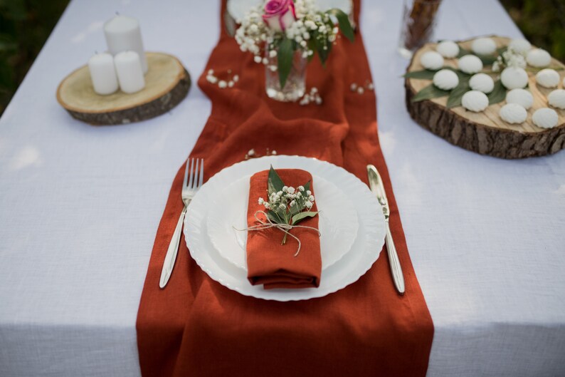 May include: A white table setting with a rust-colored linen runner. A white plate with a rust-colored napkin folded inside is set on the table. The napkin is decorated with a sprig of white flowers and green leaves. A silver fork and knife are placed on the table.