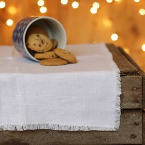 May include: A white linen table runner with fringed edges sits on a wooden table. A blue and white ceramic cup spills chocolate chip cookies onto the table runner.