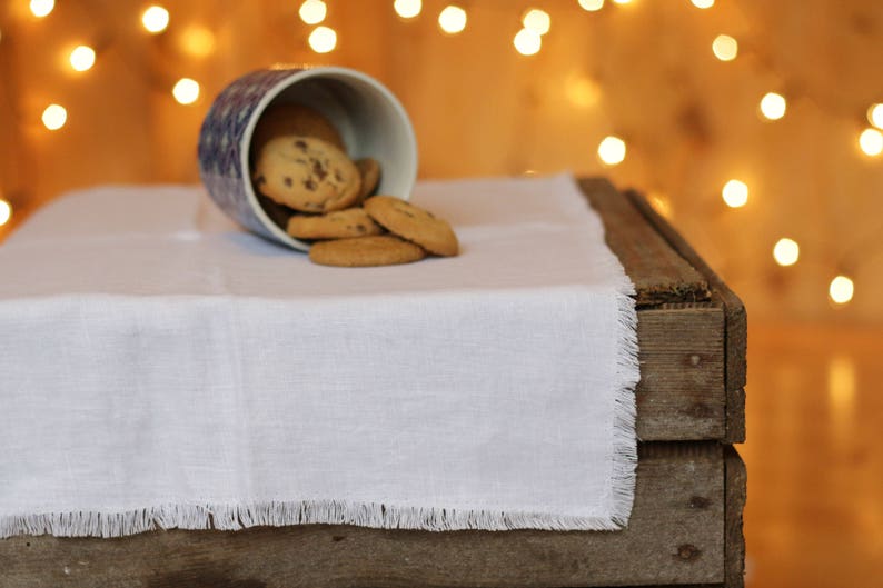 May include: A white linen table runner with fringed edges sits on a wooden table. A blue and white ceramic cup spills chocolate chip cookies onto the table runner.