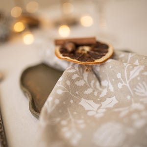 May include: A beige linen napkin with a white floral pattern, decorated with a dried orange slice and cinnamon stick. The napkin is placed on a brown tray.