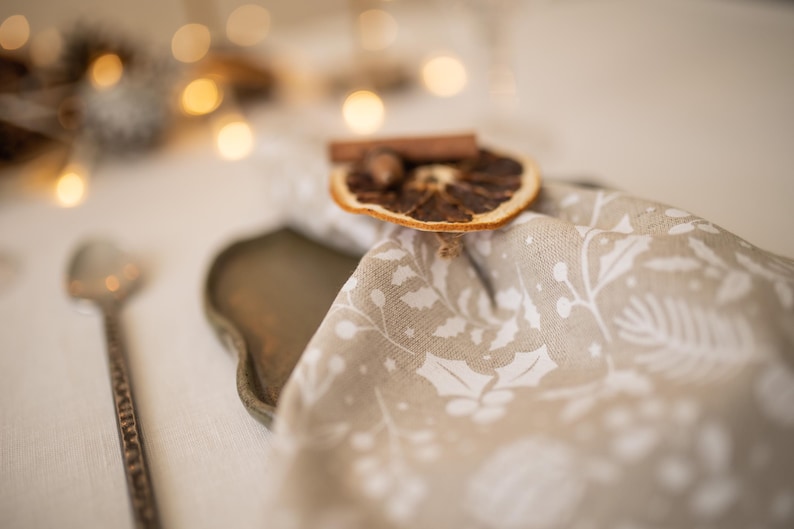 May include: A beige linen napkin with a white floral pattern, decorated with a dried orange slice and cinnamon stick. The napkin is placed on a brown tray.