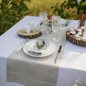 May include: A white tablecloth with a gray linen runner, set for a dinner party. The table is decorated with white candles, a small bouquet of white flowers, and a white plate with a gray napkin. There are also two wooden platters with white and pink decorations.