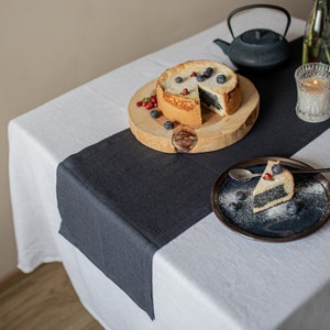 Op de afbeelding: Een zwarte linnen tafelkleed op een witte tafel met een poppyseed cake op een houten snijplank en een plak cake op een blauw bord.