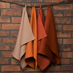 May include: Three kitchen towels hanging on a brick wall. The towels are made of linen and are in shades of beige, orange, and rust.