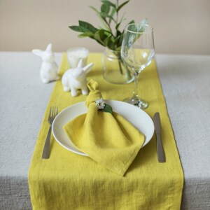 May include: A yellow linen table runner with a white napkin folded into a square and a small white flower on top. A white ceramic bunny figurine is on the table runner. A clear glass vase with green leaves is on the table runner. A clear wine glass is on the table runner. A silver fork and knife are on the table runner.