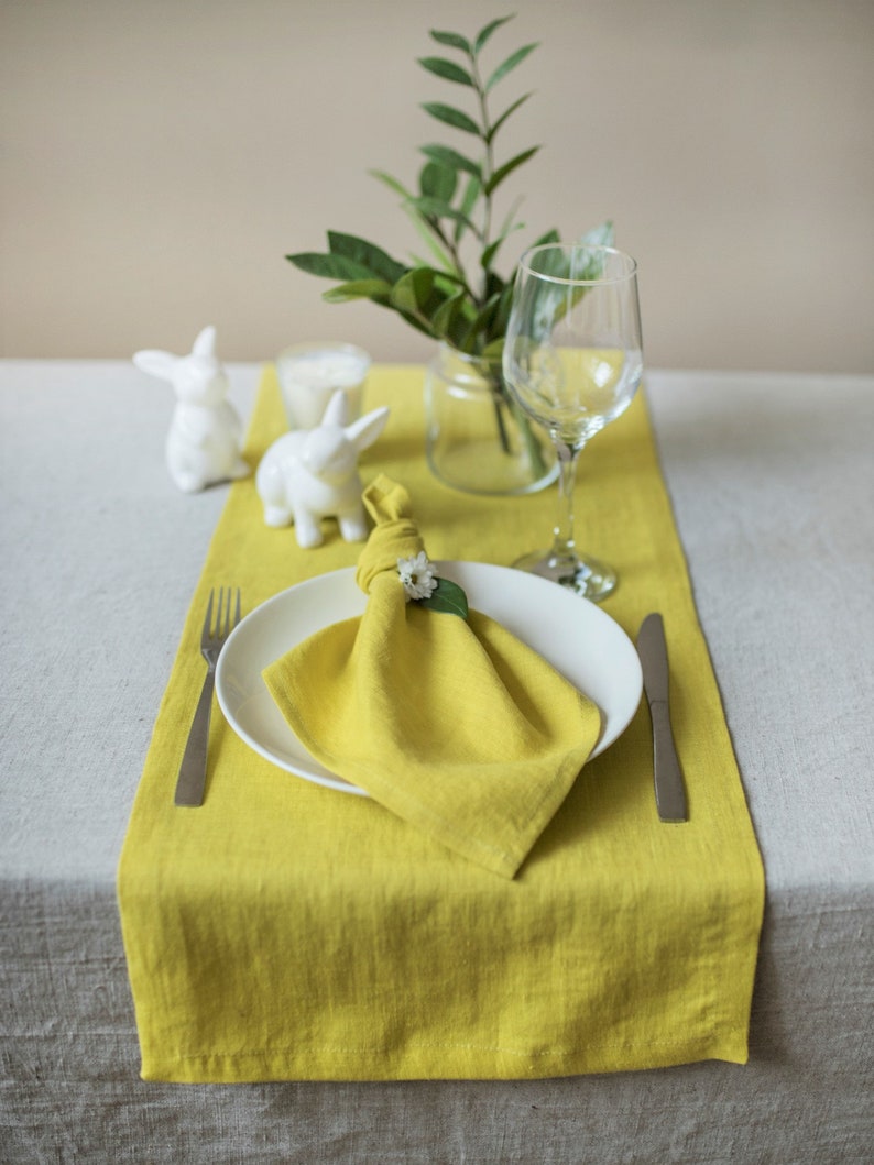 May include: A yellow linen table runner with a white napkin folded into a square and a small white flower on top. A white ceramic bunny figurine is on the table runner. A clear glass vase with green leaves is on the table runner. A clear wine glass is on the table runner. A silver fork and knife are on the table runner.