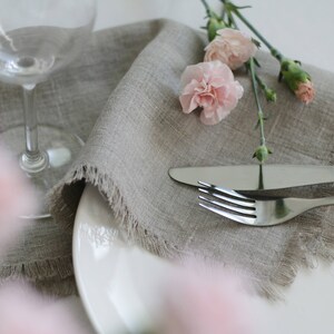 May include: A gray linen napkin with frayed edges is placed on a white plate with a fork and knife. Pink carnations are arranged on top of the napkin.
