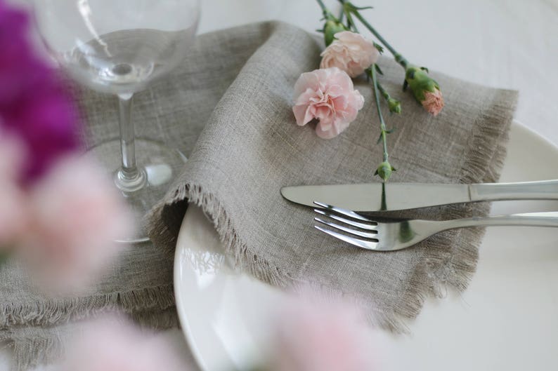 May include: A gray linen napkin with frayed edges is placed on a white plate with a fork and knife. Pink carnations are arranged on top of the napkin.