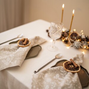 May include: A white tablecloth with two place settings. Each setting includes a white napkin with a dried orange slice and cinnamon stick as a napkin ring, a silver spoon, and a gray charger plate. The centerpiece is a gold candle holder with four lit candles and a decorative arrangement of pine cones, dried orange slices, and white berries.