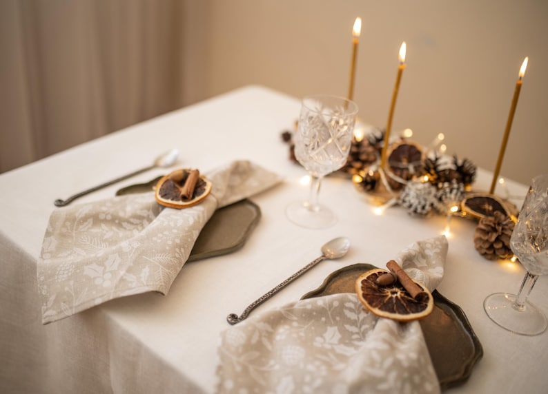 May include: A white tablecloth with two place settings. Each setting includes a white napkin with a dried orange slice and cinnamon stick as a napkin ring, a silver spoon, and a gray charger plate. The centerpiece is a gold candle holder with four lit candles and a decorative arrangement of pine cones, dried orange slices, and white berries.