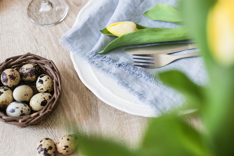Pu&ograve; includere: Un piatto bianco con un tovagliolo di lino blu, una forchetta e un tulipano giallo. Un piccolo cestino intrecciato pieno di uova di quaglia si trova su un tavolo di legno.