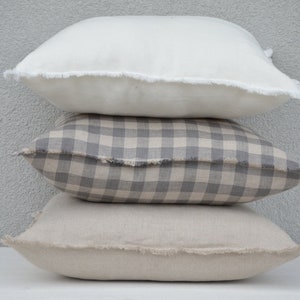May include: Three decorative throw pillows stacked on top of each other. The top pillow is white with a frayed edge. The middle pillow is a gray and white gingham pattern with a frayed edge. The bottom pillow is a light beige with a frayed edge.