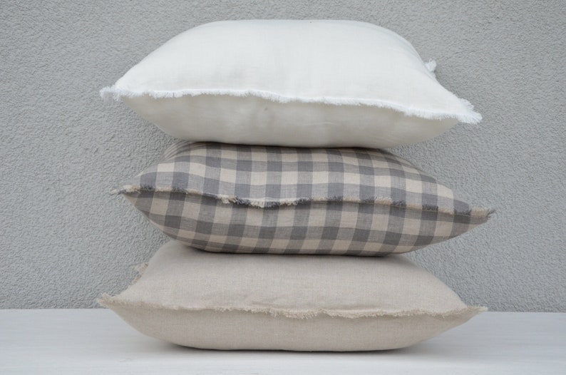 May include: Three decorative throw pillows stacked on top of each other. The top pillow is white with a frayed edge. The middle pillow is a gray and white gingham pattern with a frayed edge. The bottom pillow is a light beige with a frayed edge.