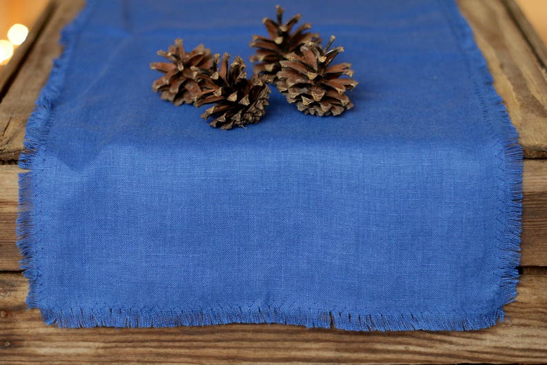 May include: A blue linen table runner with a fringed edge. Three pine cones are arranged on the runner.