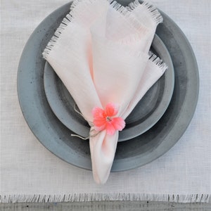 May include: A pink linen napkin with frayed edges is folded and placed on a set of gray ceramic plates. A pink flower is used as a napkin ring.