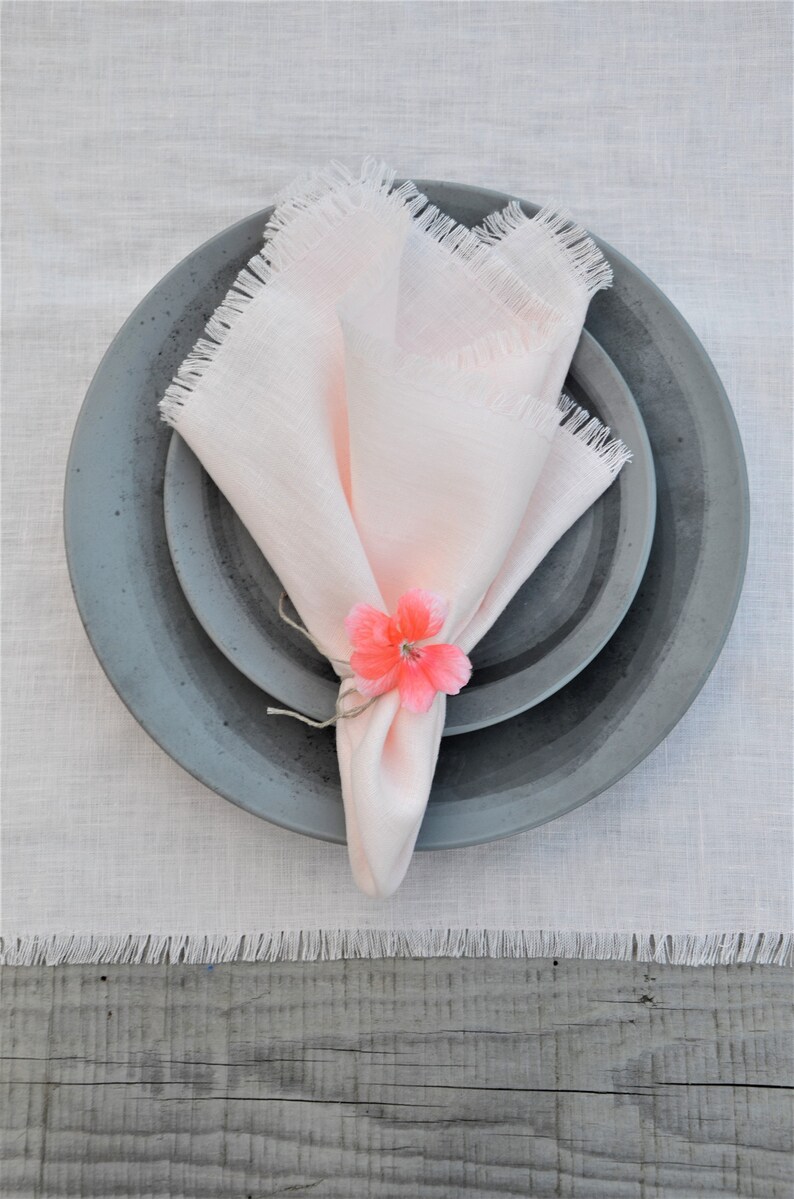 May include: A pink linen napkin with frayed edges is folded and placed on a set of gray ceramic plates. A pink flower is used as a napkin ring.