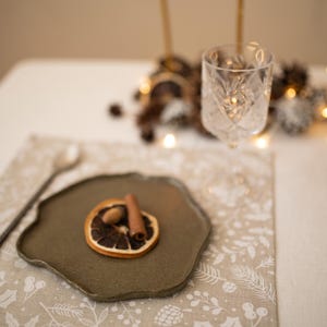 May include: A beige placemat with a white floral pattern. A small green plate with a dried orange slice, cinnamon stick, and acorn sits on the placemat. A silver fork and spoon are on the table.