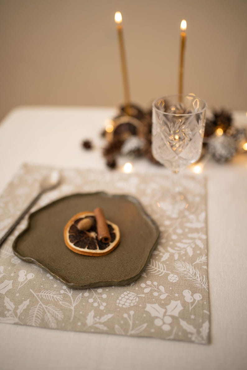 May include: A beige placemat with a white floral pattern. A small green plate with a dried orange slice, cinnamon stick, and acorn sits on the placemat. A silver fork and spoon are on the table.