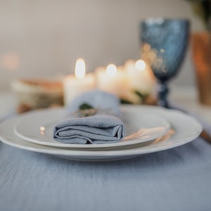 May include: A blue linen napkin folded and placed on a white plate, with a second plate underneath. The plates are on a blue linen tablecloth. The background is blurred and shows candles and a glass.