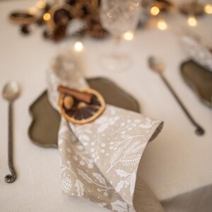 May include: A beige linen napkin with a white floral pattern is folded and placed on a brown plate. A cinnamon stick and a dried orange slice are used as a napkin ring.