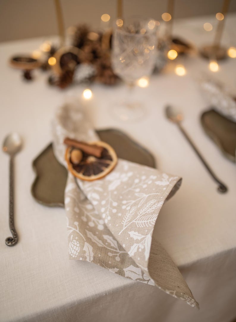 May include: A beige linen napkin with a white floral pattern is folded and placed on a brown plate. A cinnamon stick and a dried orange slice are used as a napkin ring.