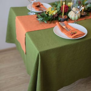 May include: A green linen tablecloth with an orange linen runner. The table is set with two place settings, each with a white plate, a folded orange linen napkin, and a fork and knife. There is a centerpiece of greenery and flowers in the middle of the table.