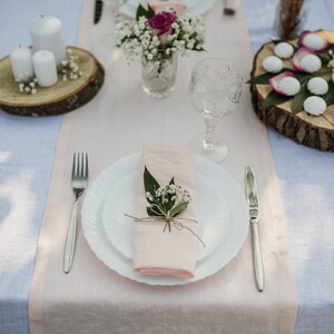 May include: A table setting with a pink linen runner, white plates, silverware, and a glass of water. There are small white flowers and a pink rose in a glass vase in the center of the table. There are also small white cookies on a wooden plate.