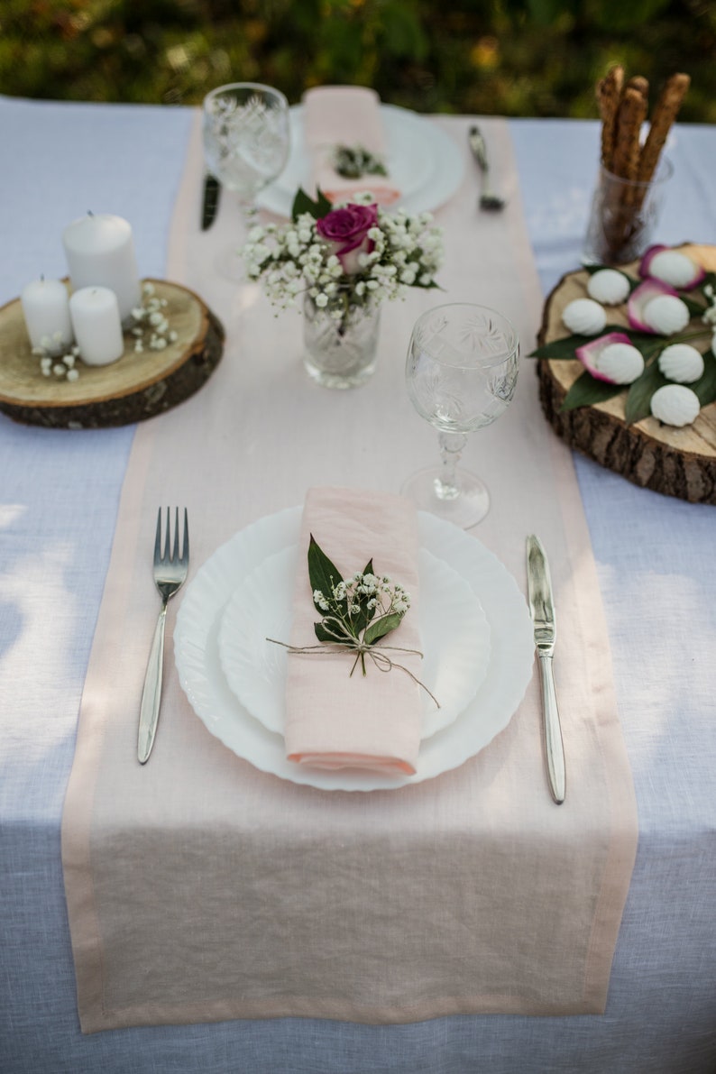 May include: A table setting with a pink linen runner, white plates, silverware, and a glass of water. There are small white flowers and a pink rose in a glass vase in the center of the table. There are also small white cookies on a wooden plate.