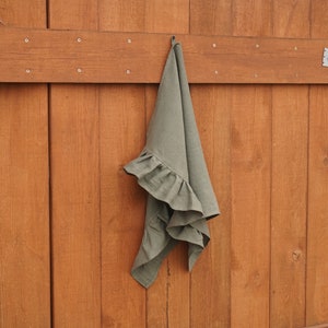 May include: A sage green linen tea towel with a ruffled edge hangs on a hook against a wood panel background.