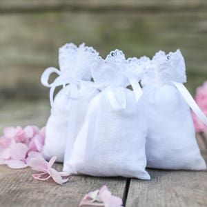 May include: Three small white linen bags with lace trim and ribbon ties are arranged on a wooden surface. Pink flower petals and a large pink flower are scattered around the bags, creating a soft, romantic scene.