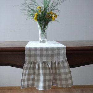 Puede incluir: Un camino de mesa a cuadros beige y blanco con un borde fruncido. El camino de mesa está sobre una mesa de madera con un jarrón de flores amarillas en el centro.