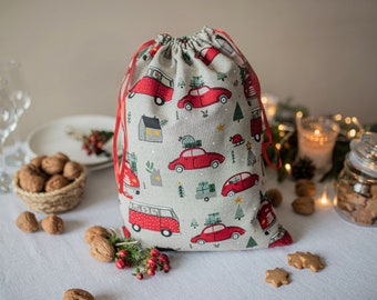 Puede incluir: Una bolsa de regalo de Navidad roja y blanca con cierre de cordón. La bolsa está hecha de tela y presenta un patrón de coches y furgonetas rojas con árboles de Navidad y casas.