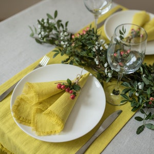 May include: A white dinner plate with a yellow linen napkin folded into a triangle and tied with a sprig of greenery and red berries. The plate is set on a yellow linen placemat with a silver fork and knife.