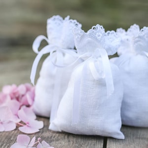 May include: Three white linen sachets with lace trim and ribbon ties, arranged on a wooden surface with pink hydrangea flowers. The sachets are filled and tied with white ribbons.