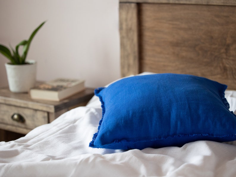 May include: A blue linen pillow with frayed edges sits on a white bed with a wooden headboard.