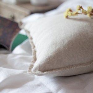 May include: A close-up of a beige linen pillow with frayed edges, resting on a white linen bedspread. Dried flowers are placed on top of the pillow.