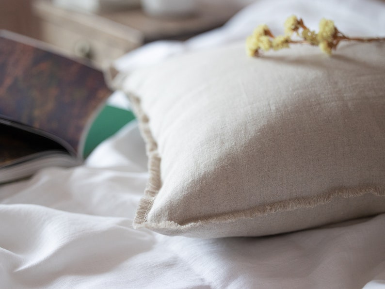May include: A close-up of a beige linen pillow with frayed edges, resting on a white linen bedspread. Dried flowers are placed on top of the pillow.