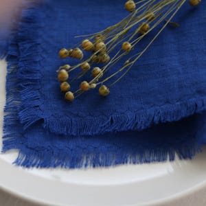 May include: A blue linen napkin with frayed edges, folded and placed on a white plate. A sprig of dried flax flowers with seed pods is placed on top of the napkin.